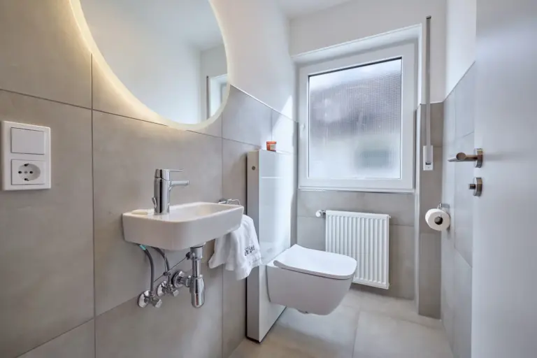 Modern half bath in CORAL BLOOM with wall-mounted toilet, compact washbasin and illuminated round mirror